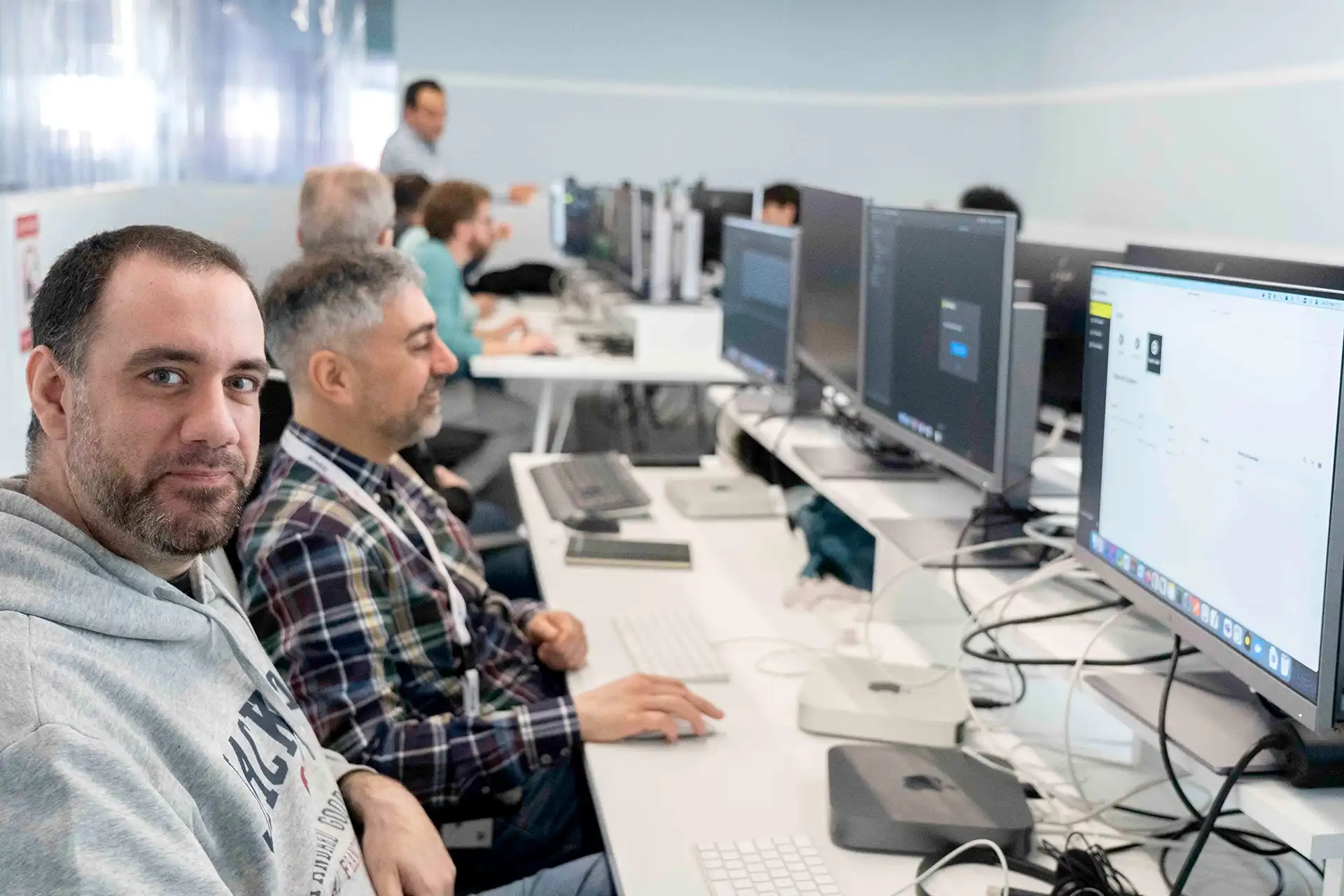 Mann in grauem Kapuzenpullover schaut in die Kamera in einem Computerlabor, während Kollegen arbeiten