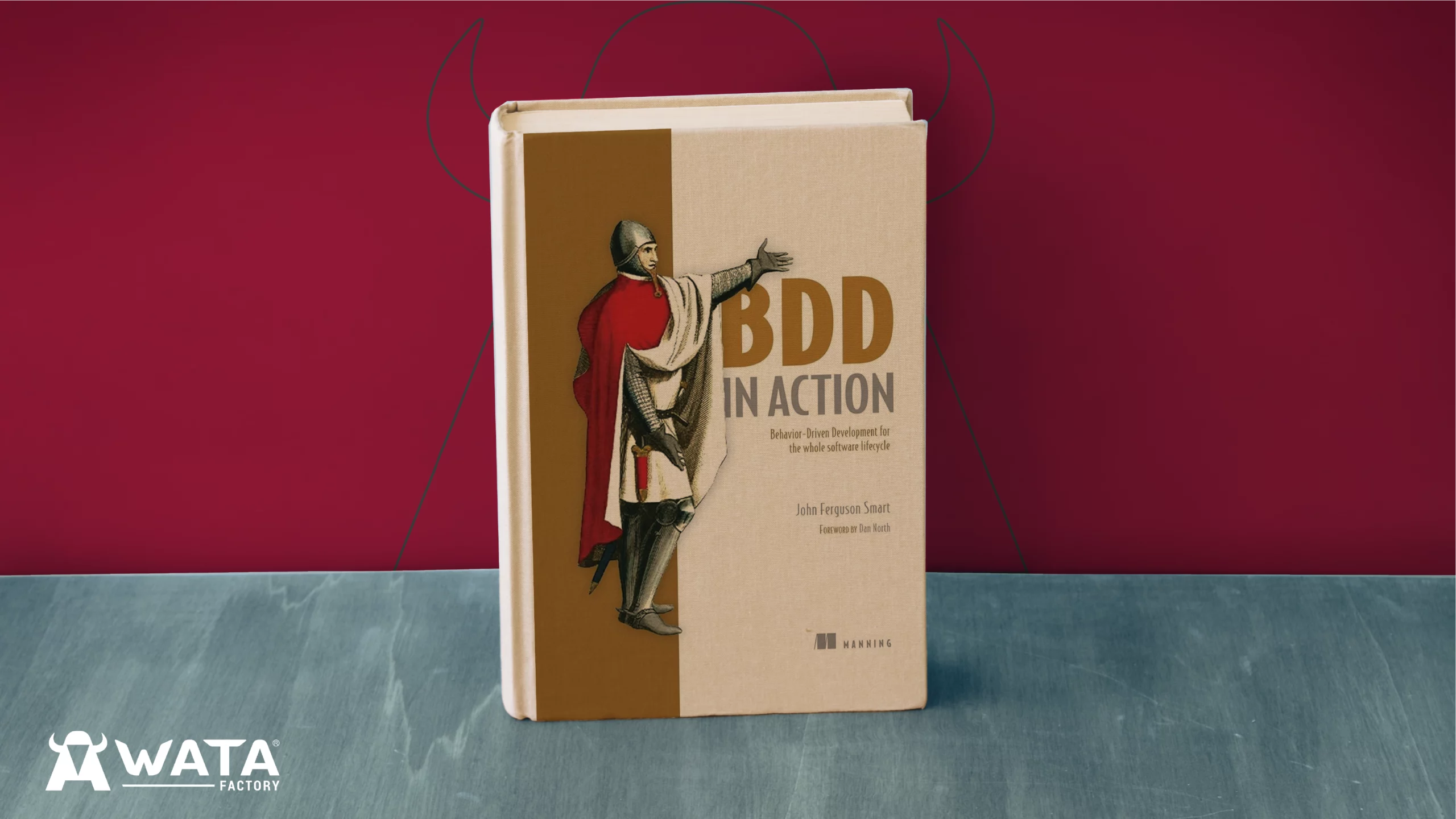 Portada del libro BDD in Action de John Ferguson Smart, colocado sobre un fondo con los colores corporativos de WATA Factory y el logo de la empresa visible detrás.