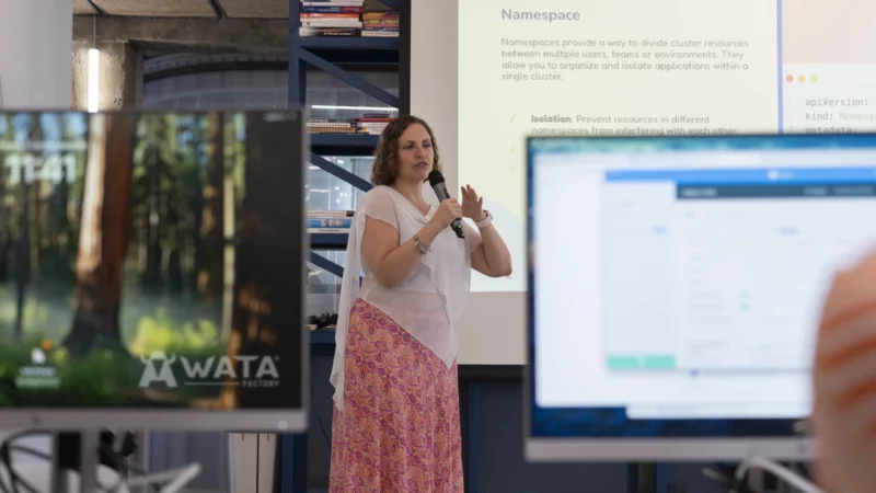 Laura Morillo-Velarde en el escenario de WATA Factory presentando sobre DevOps y Kubernetes, con diapositivas proyectadas y pantallas de compañeros en primer plano.