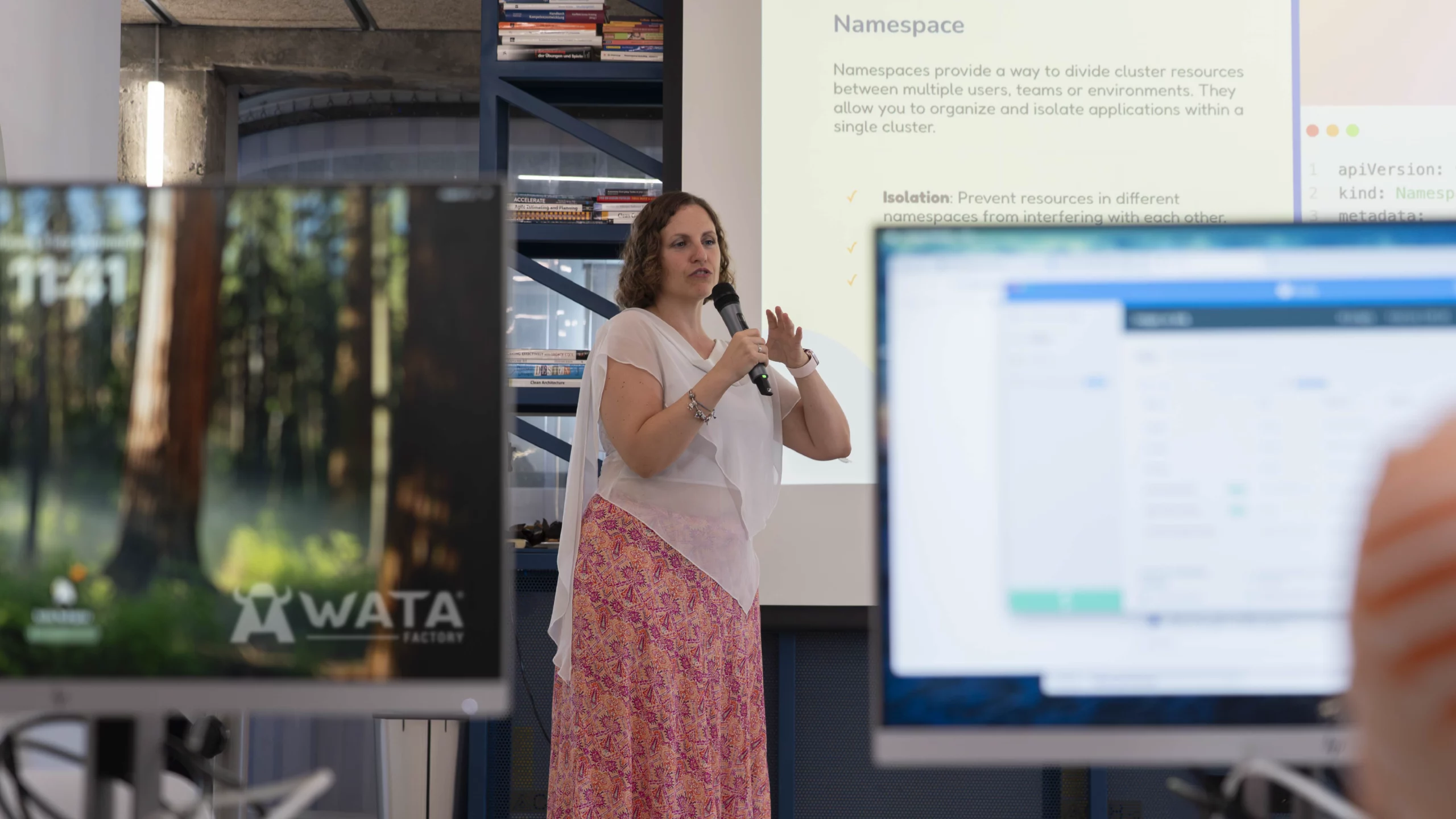 Laura Morillo-Velarde en el escenario de WATA Factory presentando sobre DevOps y Kubernetes, con diapositivas proyectadas y pantallas de compañeros en primer plano.
