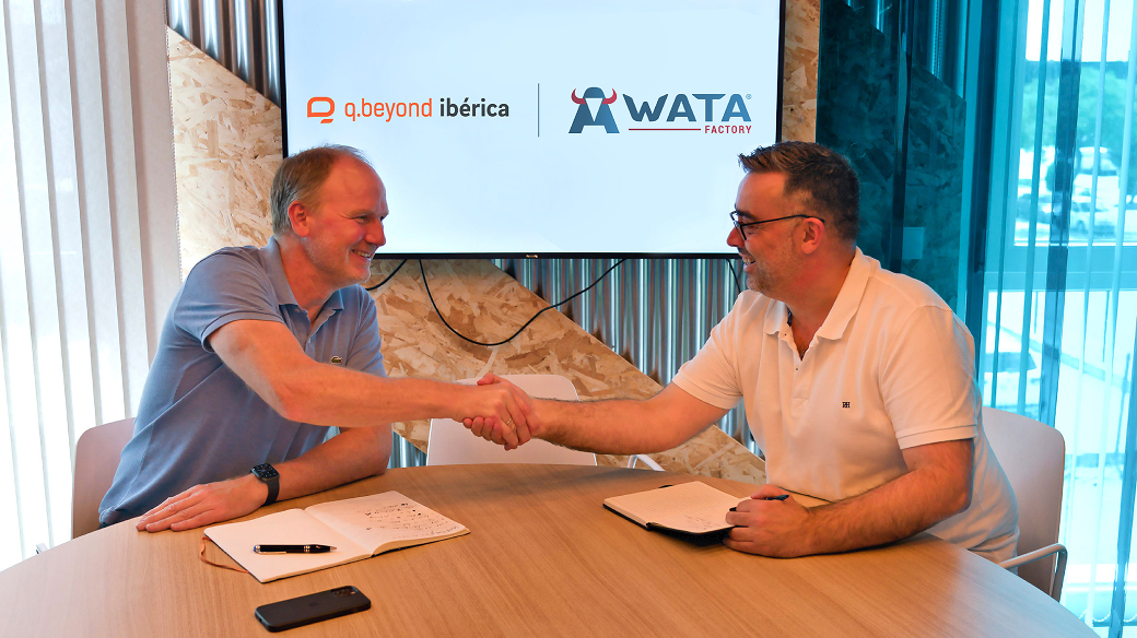 Zwei Geschäftsleute schütteln sich die Hände über einem Konferenztisch während eines Treffens zwischen q.beyond ibérica und WATA Factory, mit den Firmenlogos an der Wand und Dokumenten auf dem Tisch.