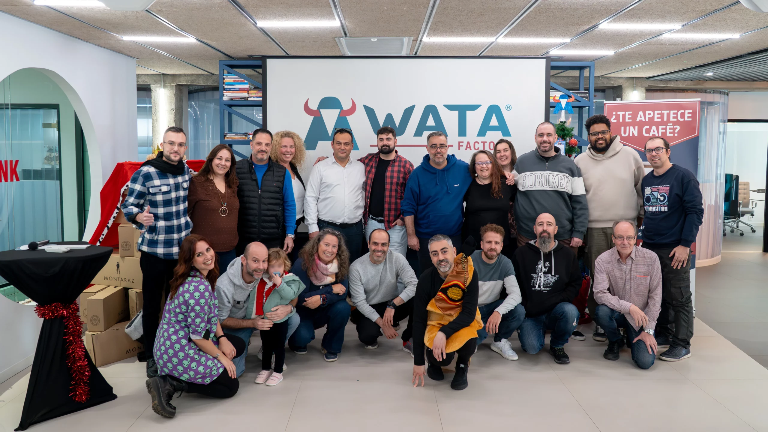 Equipo completo de WATA Factory posando junto al logo de la empresa durante el All Hands navideño, con decoración festiva y un compañero disfrazado de jamón ibérico.